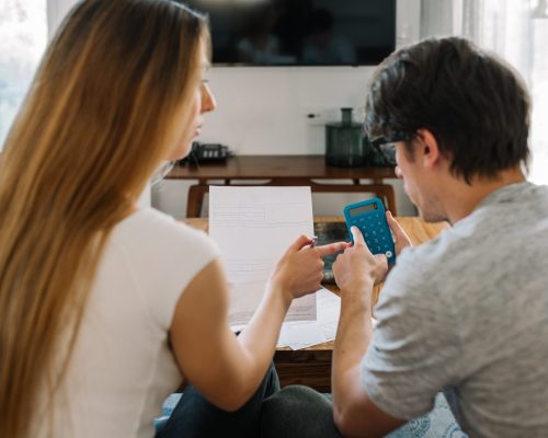 Anonymous young married couple with financial problems and emotional stress doing calculations with paper work sitting in the couch at home